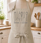 A beige kitchen apron displayed on a dress form in a bright, real kitchen. The apron features a gray line art graphic of various kitchen utensils and the text 'theresa's kitchen' printed on the chest.