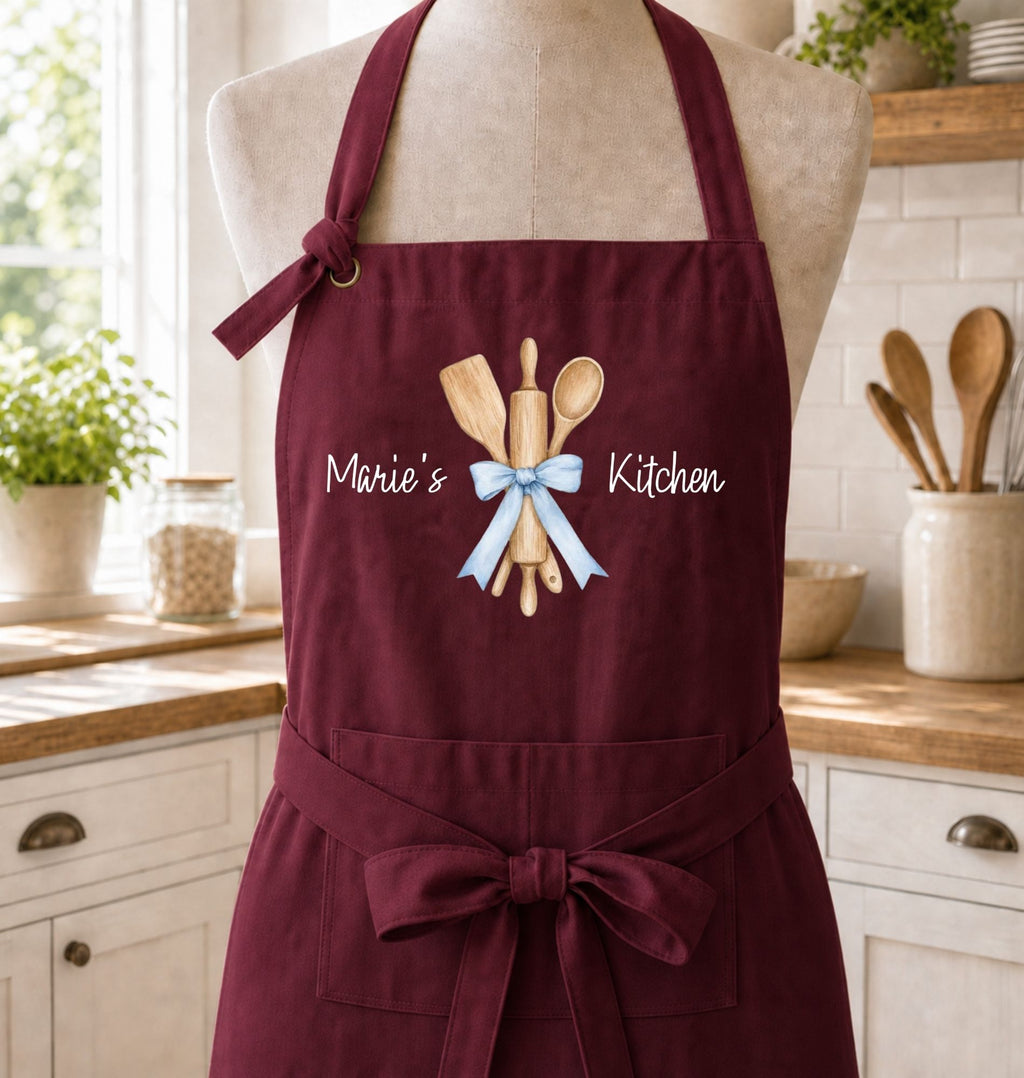 A burgundy apron displayed on a mannequin torso in a bright kitchen setting. The apron features a graphic of wooden kitchen utensils tied with a light blue ribbon and the text 'Marie's Kitchen' printed in white.