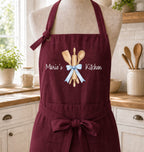A burgundy apron displayed on a mannequin torso in a bright kitchen setting. The apron features a graphic of wooden kitchen utensils tied with a light blue ribbon and the text 'Marie's Kitchen' printed in white.