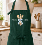 A dark green kitchen apron displayed on a mannequin torso in a bright kitchen setting. The apron features a graphic of wooden kitchen utensils tied with a light blue ribbon, with the text 'Marie's Kitchen' printed in white script.