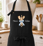 A black kitchen apron displayed on a mannequin torso in a bright kitchen setting. The apron has a front tie belt and a printed design of wooden kitchen utensils tied with a light blue ribbon, with the text 'Marie's Kitchen' in white script.