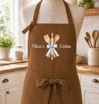 A brown kitchen apron displayed on a mannequin torso in a bright kitchen setting. The apron has a central graphic of wooden kitchen utensils tied with a light blue ribbon, with the text 'Marie's Kitchen' printed in white script.