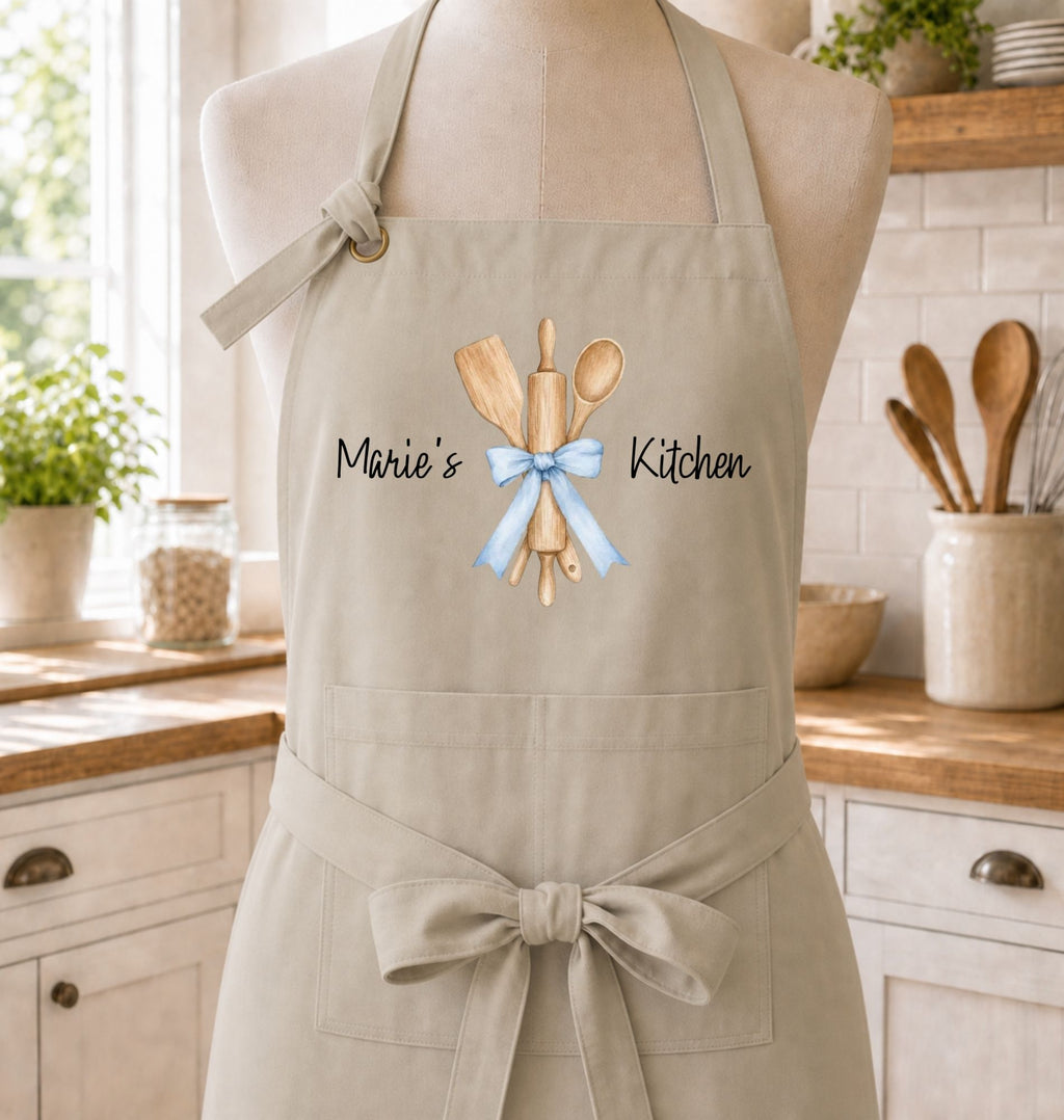 A beige kitchen apron displayed on a mannequin torso in a bright kitchen setting. The apron has a watercolor-style graphic of wooden kitchen utensils tied with a light blue ribbon, with the words 'Marie's Kitchen' printed in black script.