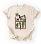 Flat lay of a Natural t-shirt featuring illustrated cats sitting and playing on a shelf of potted plants on a white background