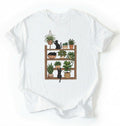 Flat lay of a white t-shirt featuring illustrated cats sitting and playing on a shelf of potted plants on a white background