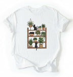 Flat lay of a white t-shirt featuring illustrated cats sitting and playing on a shelf of potted plants on a white background