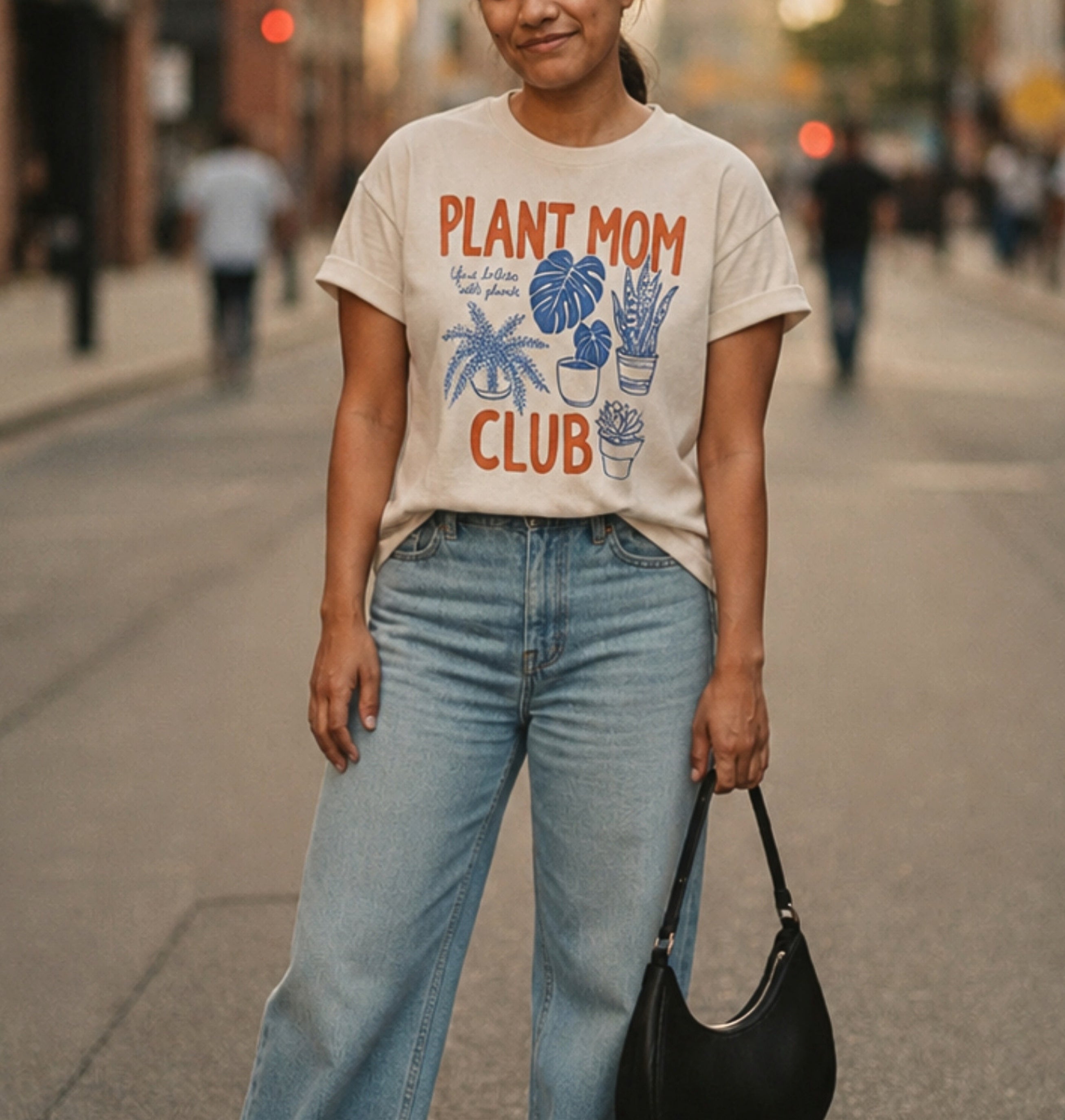 A woman standing on a city street wearing a cream graphic T-shirt that reads "PLANT MOM CLUB" with blue plant illustrations, paired with light blue jeans and holding a black handbag.