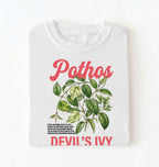 Flat lay of a white t-shirt featuring a botanical leaf illustration of Pothos leaves with text reading “Pothos Devil’s Ivy” perfect gift for plant lovers and houseplant enthusiasts on white background 