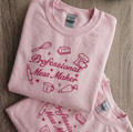 A folded light pink t-shirt with a dark pink graphic print that reads 'Professional Mess Maker' surrounded by baking-themed illustrations, displayed on a wooden surface.