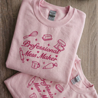 A folded light pink t-shirt with a dark pink graphic print that reads 'Professional Mess Maker' surrounded by baking-themed illustrations, displayed on a wooden surface.