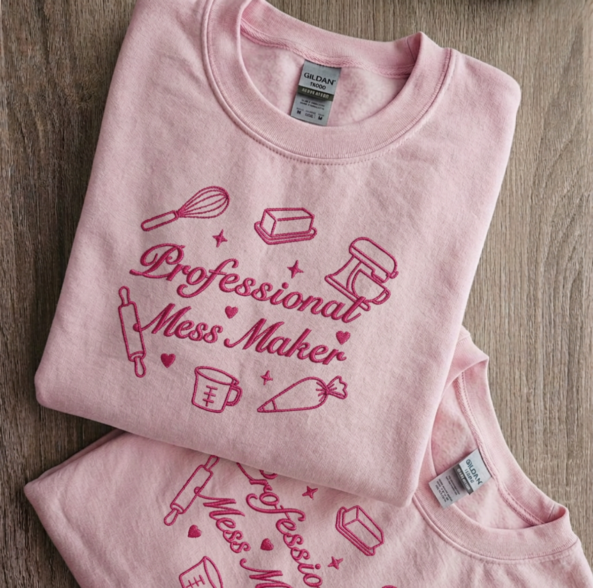 A folded light pink t-shirt with a dark pink graphic print that reads 'Professional Mess Maker' surrounded by baking-themed illustrations, displayed on a wooden surface.