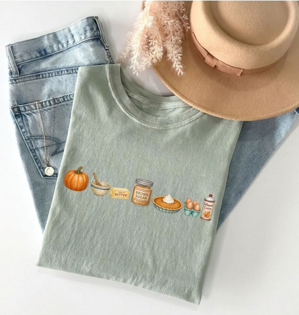 A folded sage green t-shirt with a colorful autumn-themed graphic print, styled in a flat-lay with light-wash jeans, a tan hat, and a necklace on a white background.