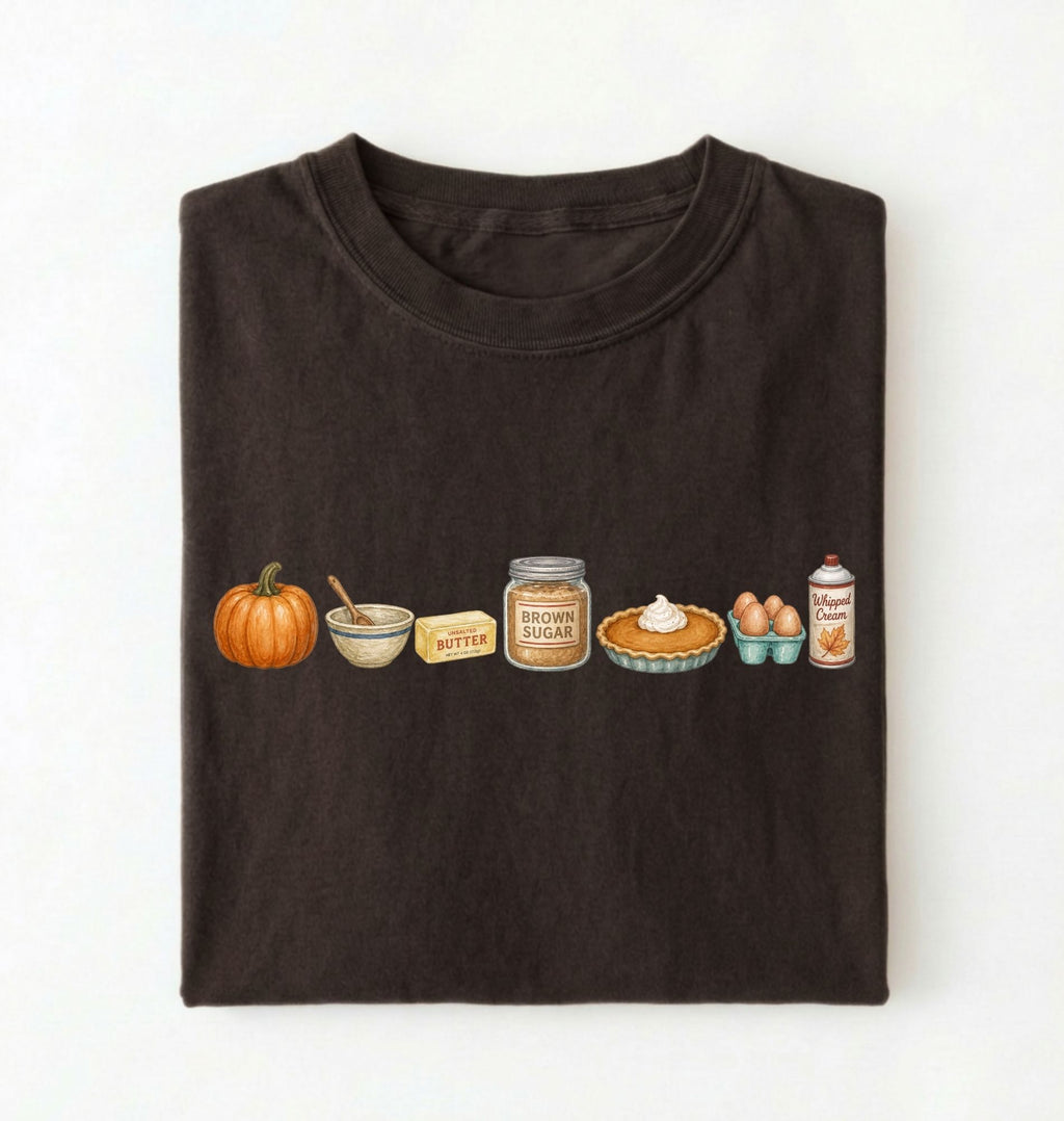 A dark brown folded t-shirt on a white background, featuring a horizontal graphic print of autumn baking ingredients including a pumpkin, butter, brown sugar, pie, eggs, and a can of whipped cream.