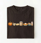 A dark brown folded t-shirt on a white background, featuring a horizontal graphic print of autumn baking ingredients including a pumpkin, butter, brown sugar, pie, eggs, and a can of whipped cream.