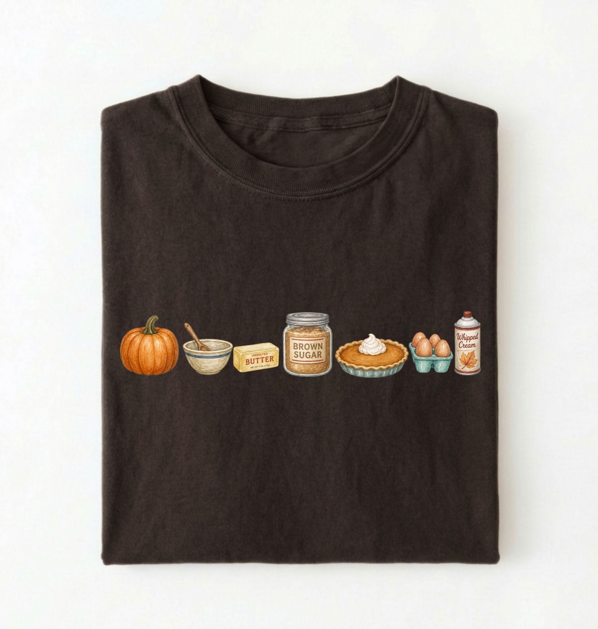 A dark brown folded t-shirt on a white background, featuring a horizontal graphic print of autumn baking ingredients including a pumpkin, butter, brown sugar, pie, eggs, and a can of whipped cream.