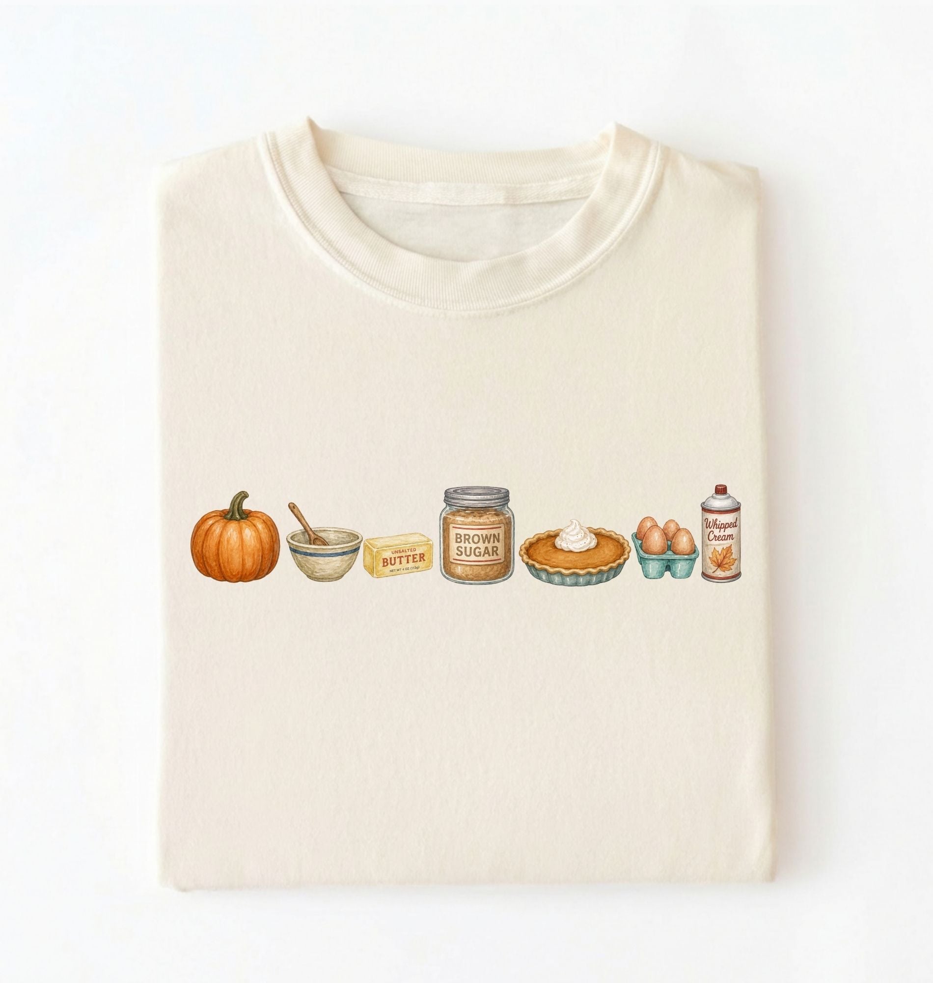 A folded cream-colored t-shirt on a white background, featuring a horizontal watercolor-style graphic of autumn baking ingredients including a pumpkin, butter, brown sugar, pie, eggs, and vanilla.