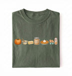 An olive green folded t-shirt with a horizontal graphic print of autumn baking ingredients, including a pumpkin, butter, brown sugar, pie, eggs, and whipped cream, on a white background.