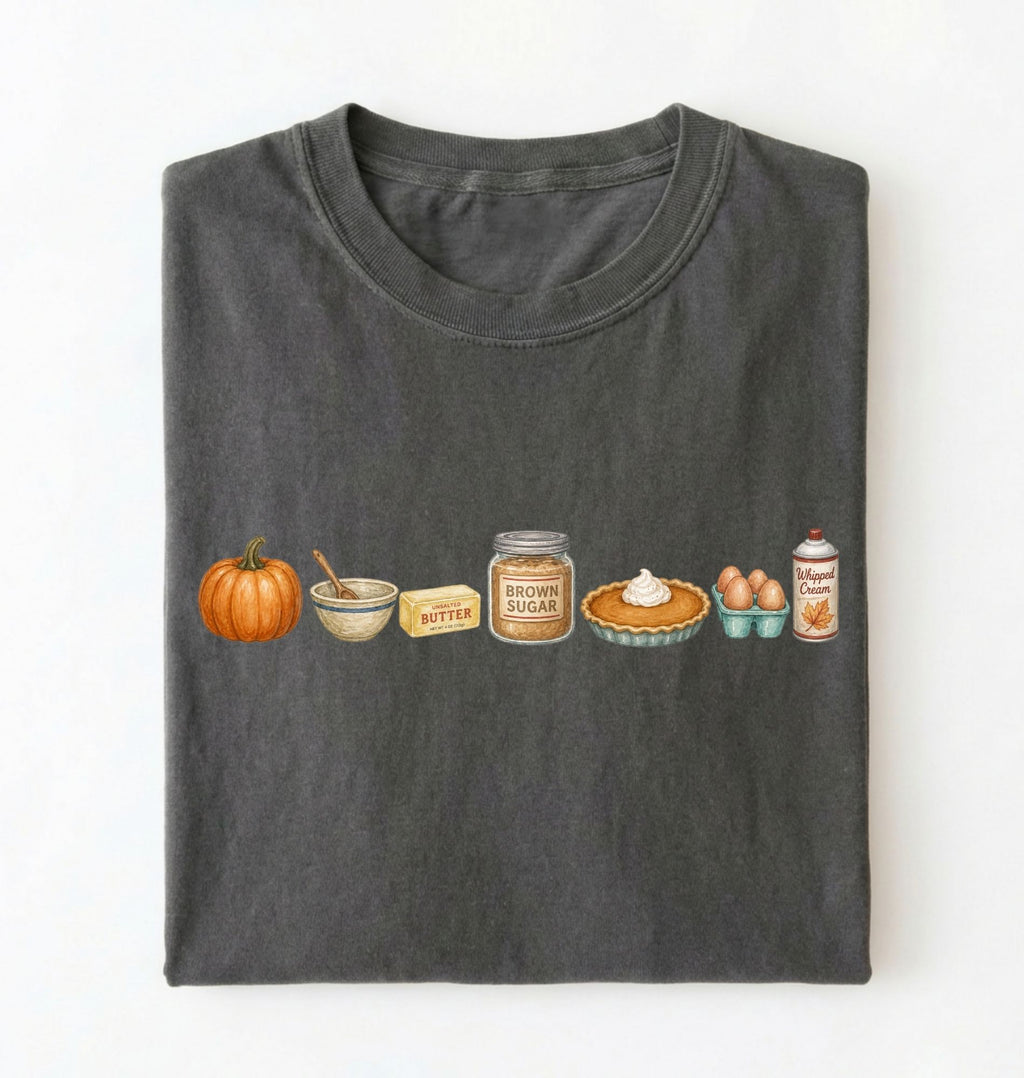 A dark gray folded t-shirt on a white background, featuring a horizontal graphic print of autumn baking ingredients including a pumpkin, butter, brown sugar, a pie, eggs, and cream.