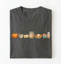 A dark gray folded t-shirt on a white background, featuring a horizontal graphic print of autumn baking ingredients including a pumpkin, butter, brown sugar, a pie, eggs, and cream.
