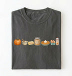 A dark gray folded t-shirt on a white background, featuring a horizontal graphic print of autumn baking ingredients including a pumpkin, butter, brown sugar, a pie, eggs, and cream.