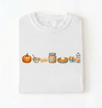 A white crewneck t-shirt laid flat on a white background, featuring a horizontal graphic illustration of autumn baking ingredients and treats, including a pumpkin, butter, brown sugar, pumpkin pie, eggs, and whipped cream.