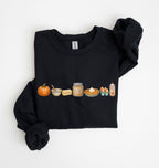 A black long-sleeve sweatshirt laid flat on a white background, featuring a horizontal graphic print of autumn-themed food items including a pumpkin, butter, cinnamon, pie, eggs, and whipped cream.