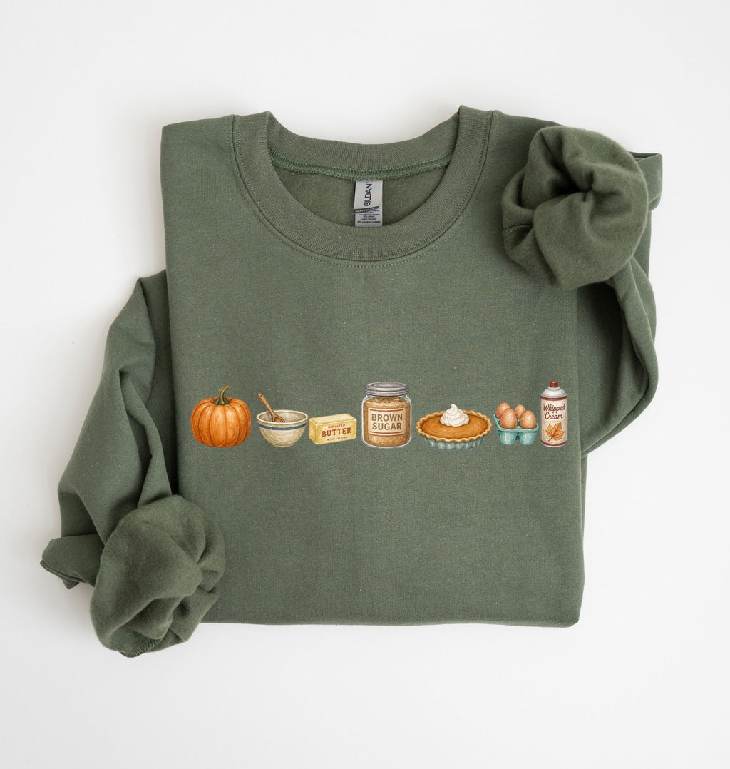 An olive green long-sleeve t-shirt with rolled cuffs, featuring a horizontal graphic print of autumn baking ingredients including a pumpkin, butter, sugar, pie, eggs, and whipped cream. The shirt is styled with a matching fabric scrunchie on a clean white background.