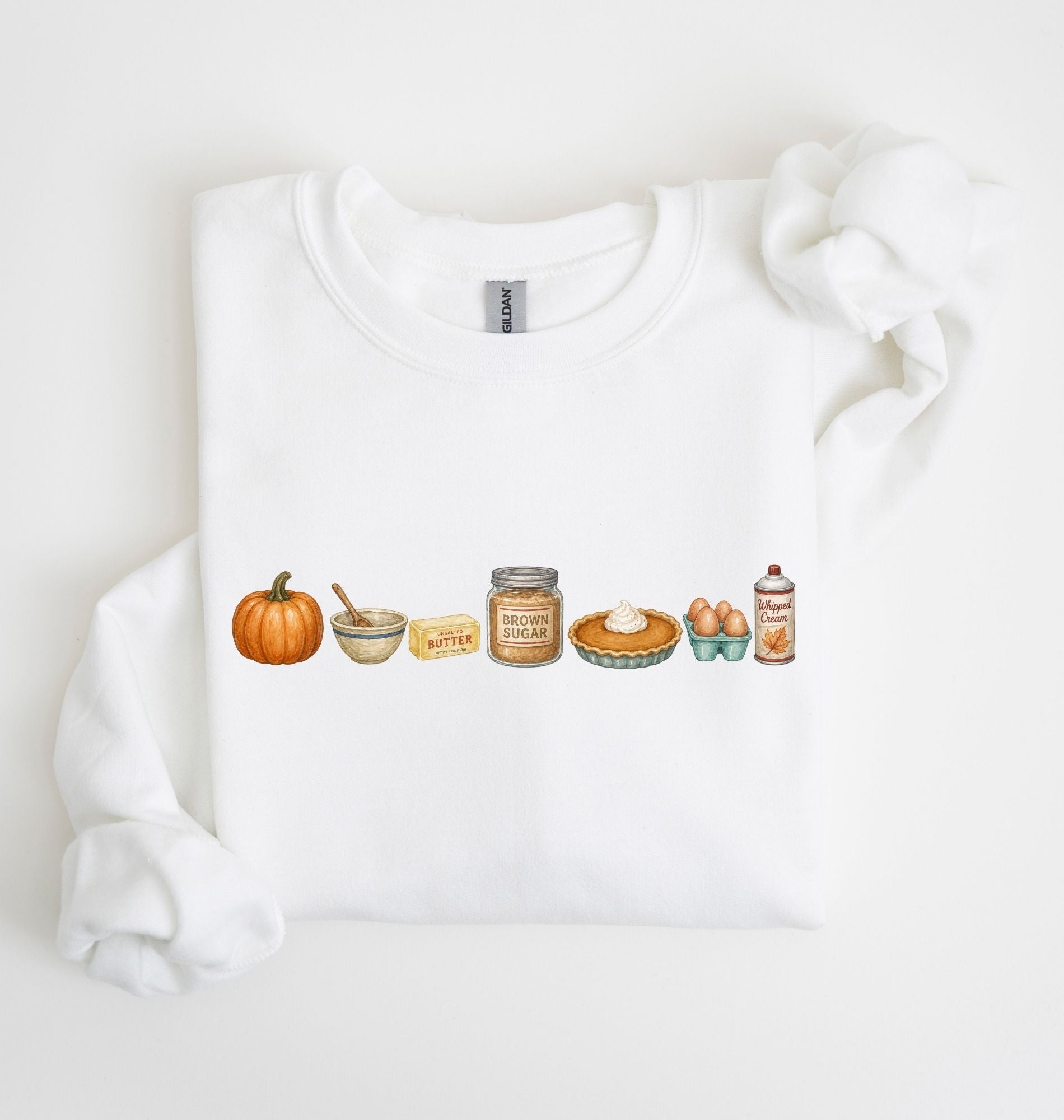 A white crewneck sweatshirt with a horizontal watercolor graphic of autumn baking elements, including a pumpkin, butter, brown sugar, pie, eggs, and cinnamon, displayed on a clean white background.