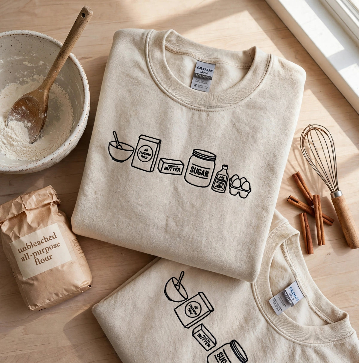 A folded cream-colored crewneck t-shirt with a black line art graphic of baking ingredients (flour, sugar, butter, eggs) on the front, styled on a light wood table with baking props like a whisk, cinnamon sticks, a bowl of flour, and a paper bag of flour.
