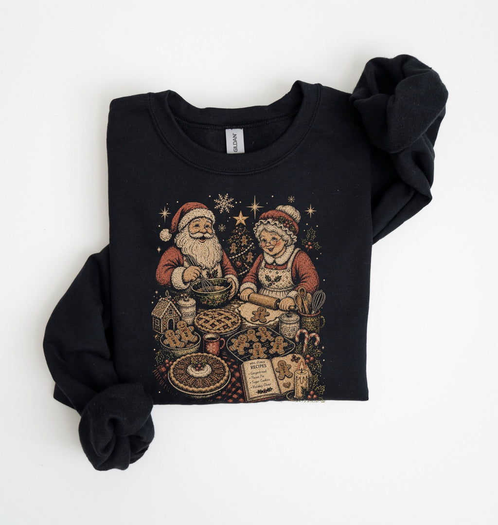 A black crewneck sweatshirt featuring a vintage-style Christmas graphic of Santa Claus and a woman baking cookies, with the sleeves rolled up, laid flat on a white background.