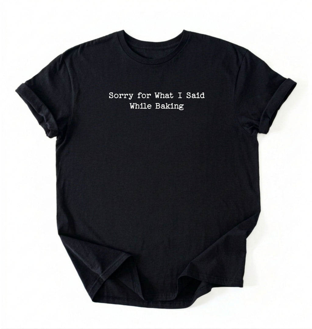 A folded black-colored t-shirt with black typewriter-style text reading 'Sorry for What I Said While Baking' on the front, displayed against a clean white background.