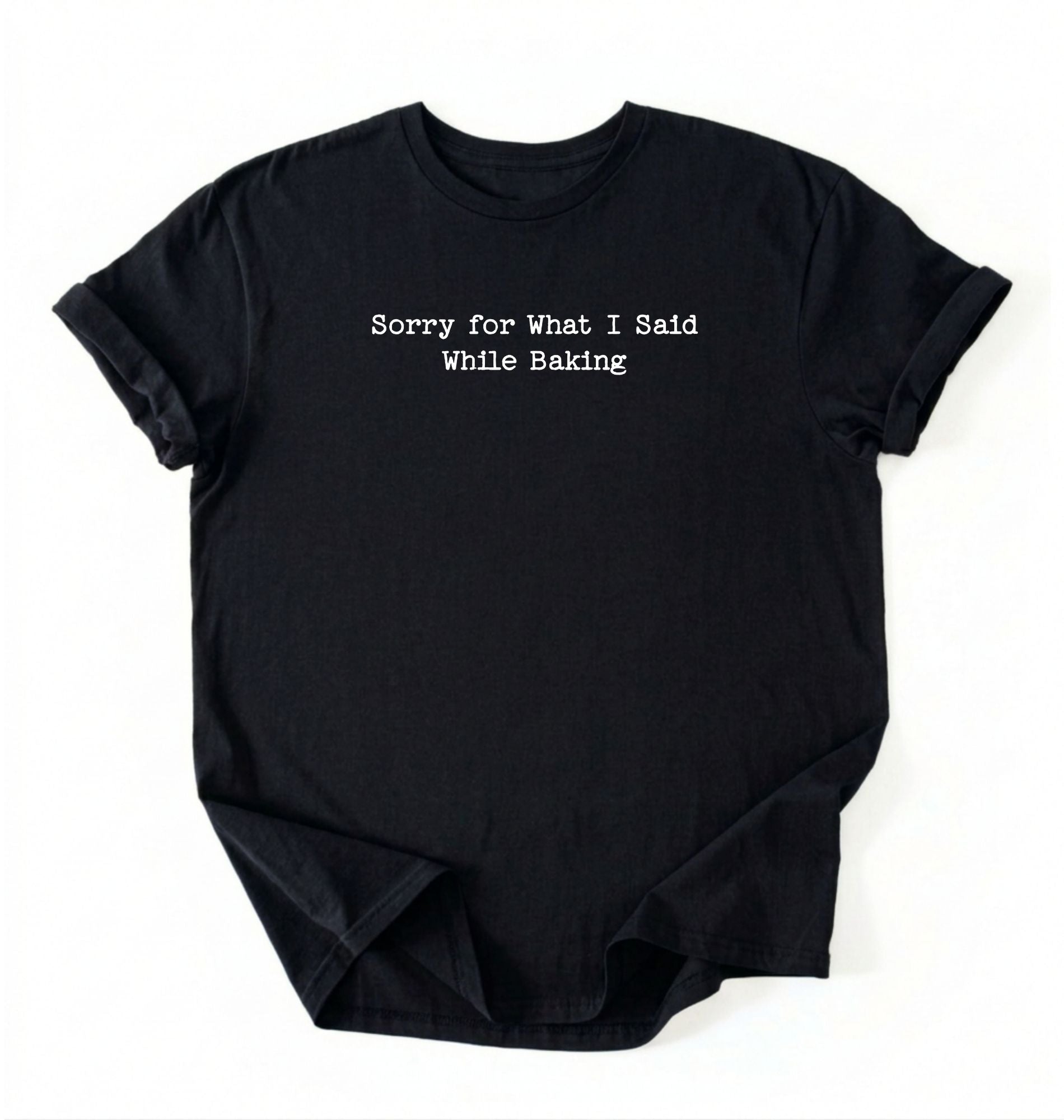 A folded black-colored t-shirt with black typewriter-style text reading 'Sorry for What I Said While Baking' on the front, displayed against a clean white background.