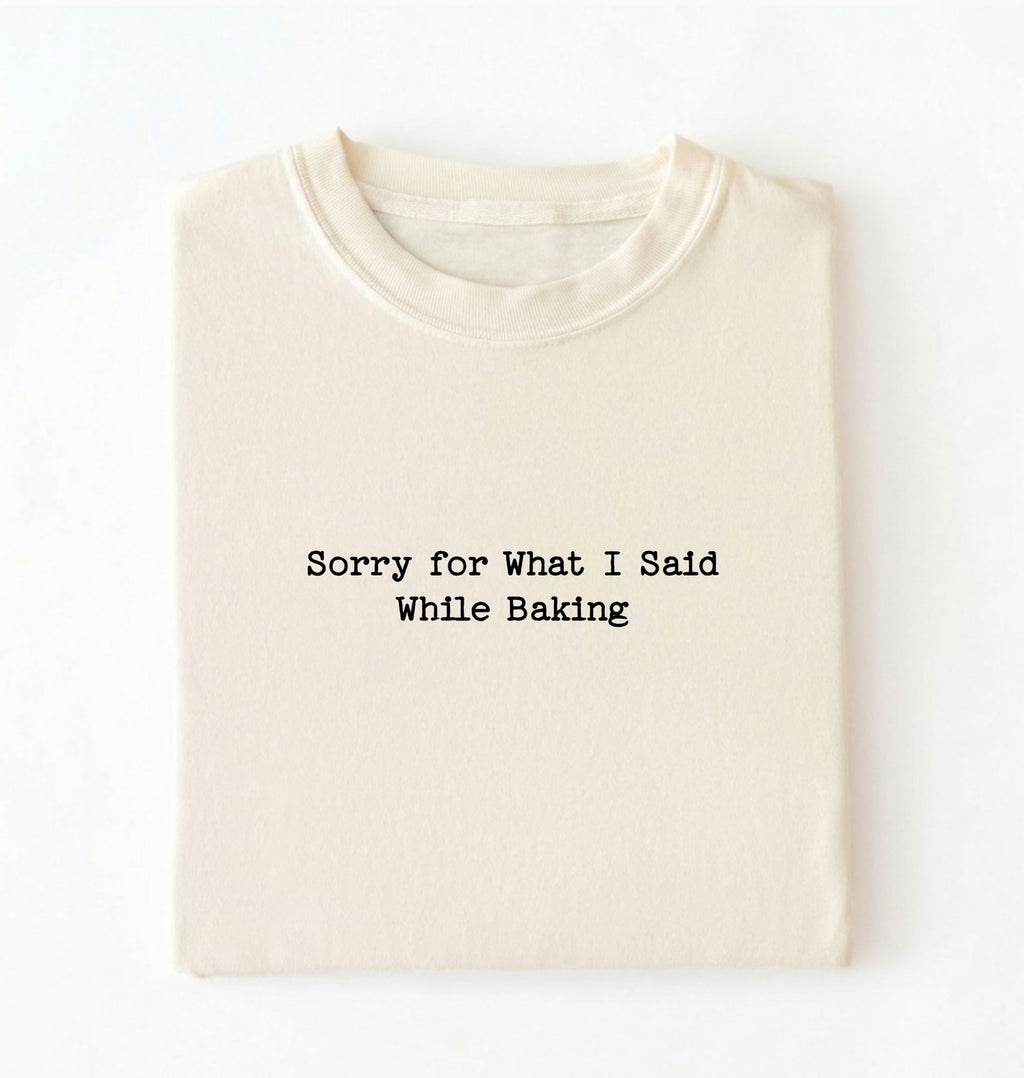 A folded cream-colored t-shirt with black typewriter-style text reading 'Sorry for What I Said While Baking' on the front, displayed against a clean white background.