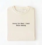 A folded cream-colored t-shirt with black typewriter-style text reading 'Sorry for What I Said While Baking' on the front, displayed against a clean white background.