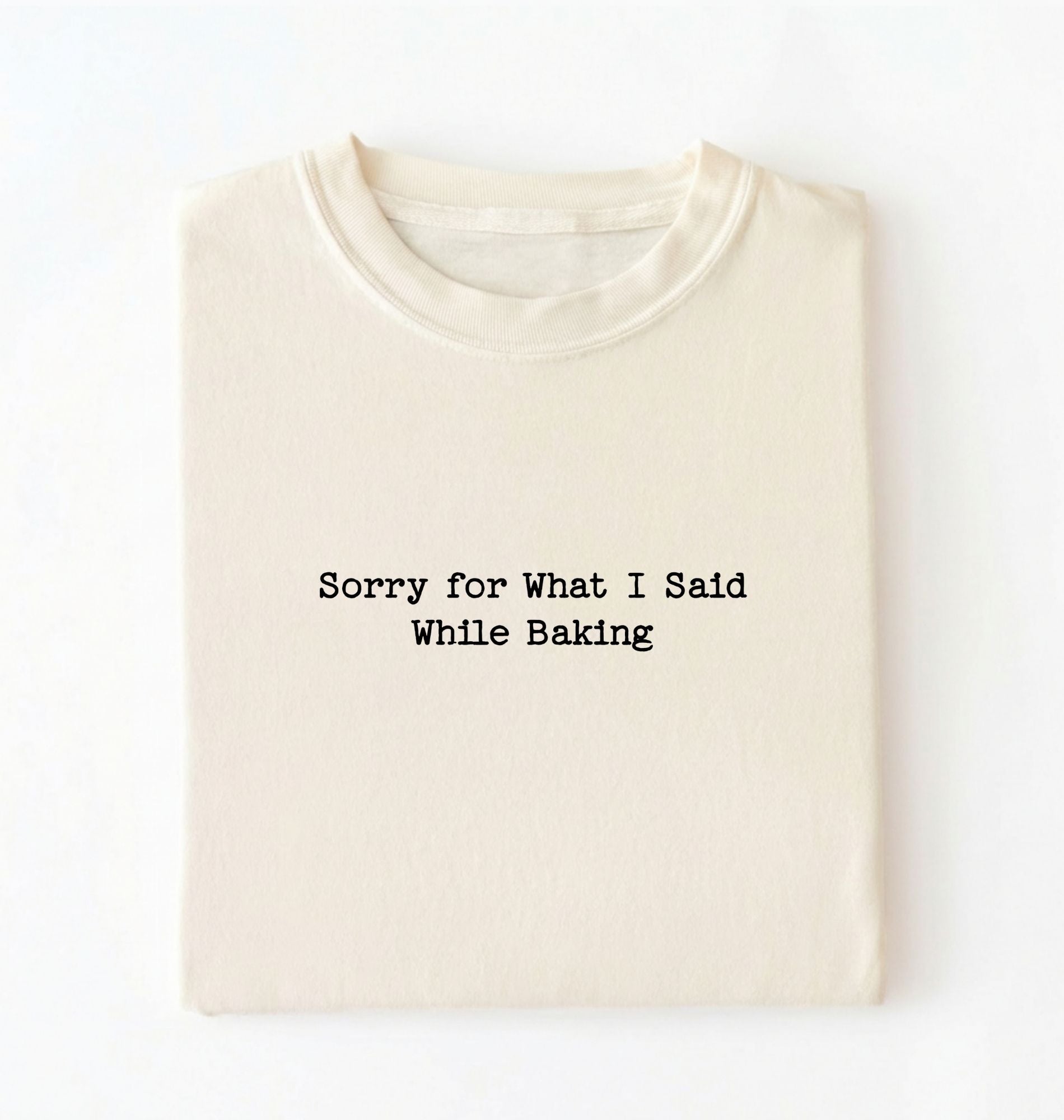 A folded cream-colored t-shirt with black typewriter-style text reading 'Sorry for What I Said While Baking' on the front, displayed against a clean white background.