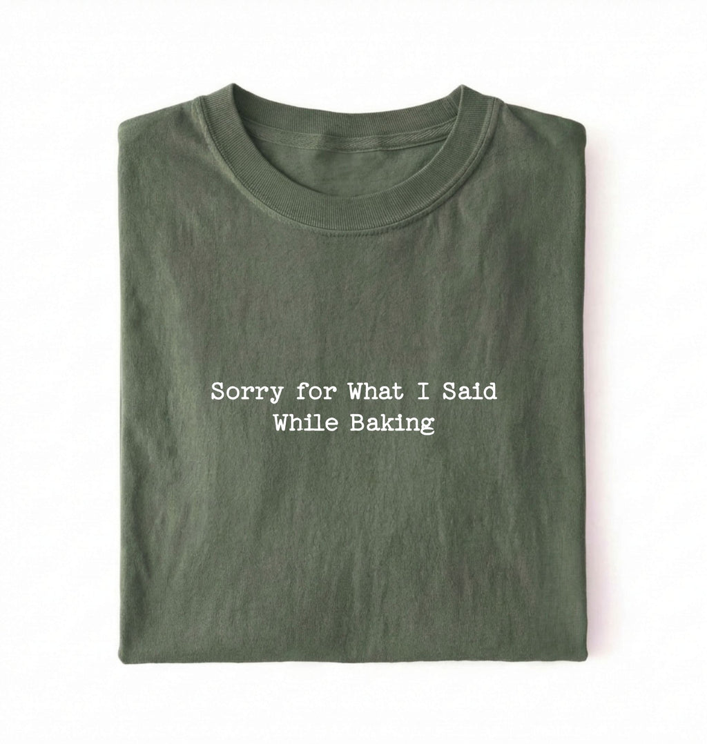 A folded green t-shirt with the black printed text 'Sorry for What I Said While Baking' centered on the chest, presented on a clean white background.