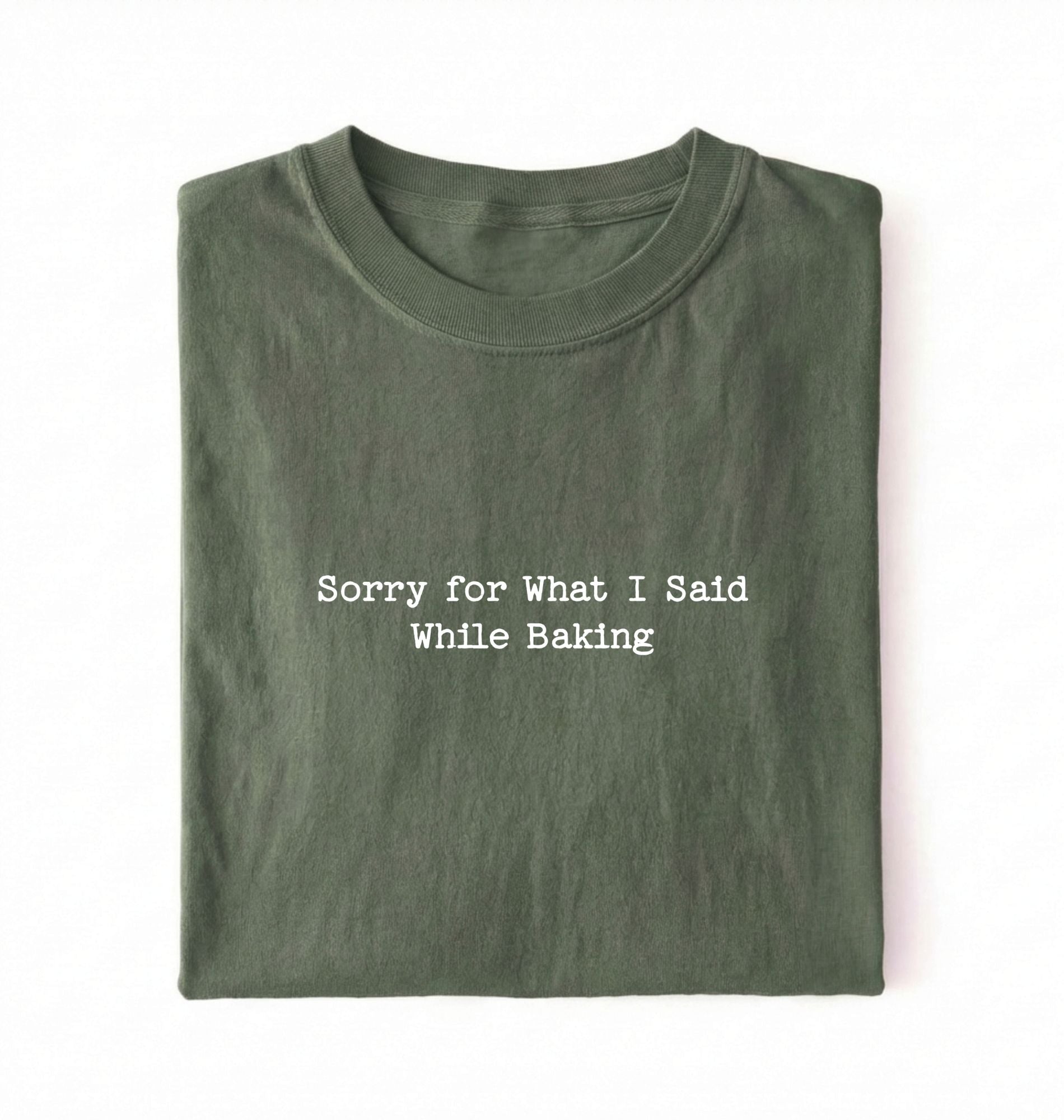 A folded green t-shirt with the black printed text 'Sorry for What I Said While Baking' centered on the chest, presented on a clean white background.