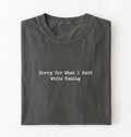 A folded pepper-colored t-shirt with black typewriter-style text reading 'Sorry for What I Said While Baking' on the front, displayed against a clean white background.