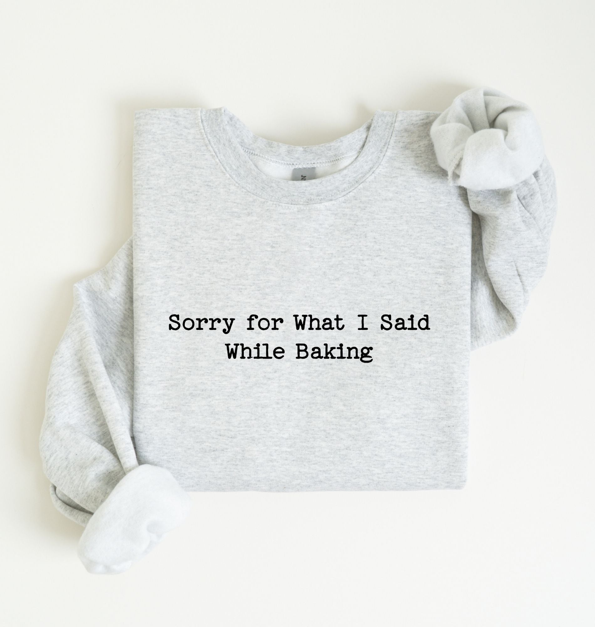 A light heather gray long-sleeve sweatshirt with the text 'Sorry for What I Said While Baking' printed in black on the front, styled flat on a light cream background.