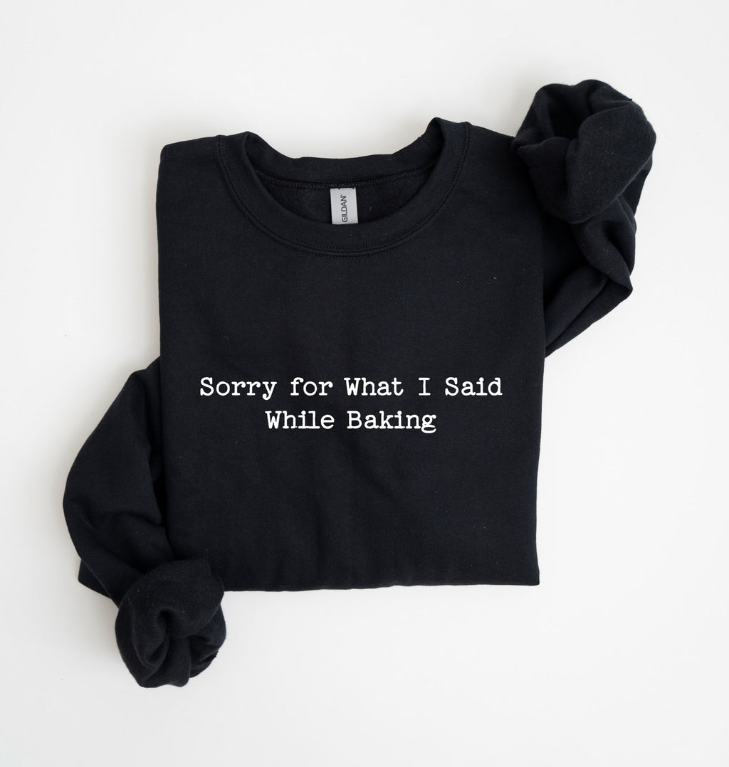 A black long-sleeve sweatshirt with the phrase 'Sorry for What I Said While Baking' printed in white typewriter-style text across the chest, shown folded on a white background.