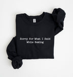 A black long-sleeve sweatshirt with the phrase 'Sorry for What I Said While Baking' printed in white typewriter-style text across the chest, shown folded on a white background.