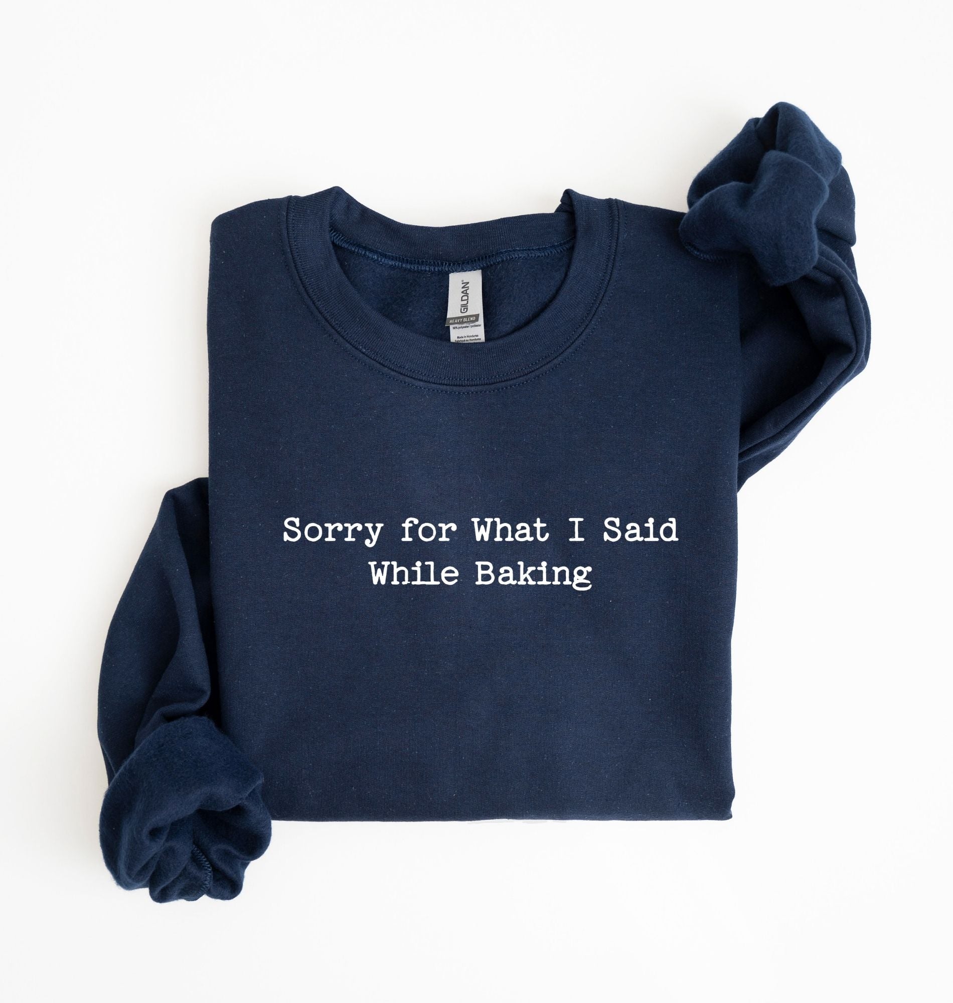 A navy blue long-sleeve sweatshirt with white text reading 'Sorry for What I Said While Baking' on the front, laid flat on a white background with the sleeves rolled up.