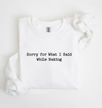 A folded white long-sleeve sweatshirt with the text 'Sorry for What I Said While Baking' printed in black typewriter-style font on the chest, photographed on a clean white background.