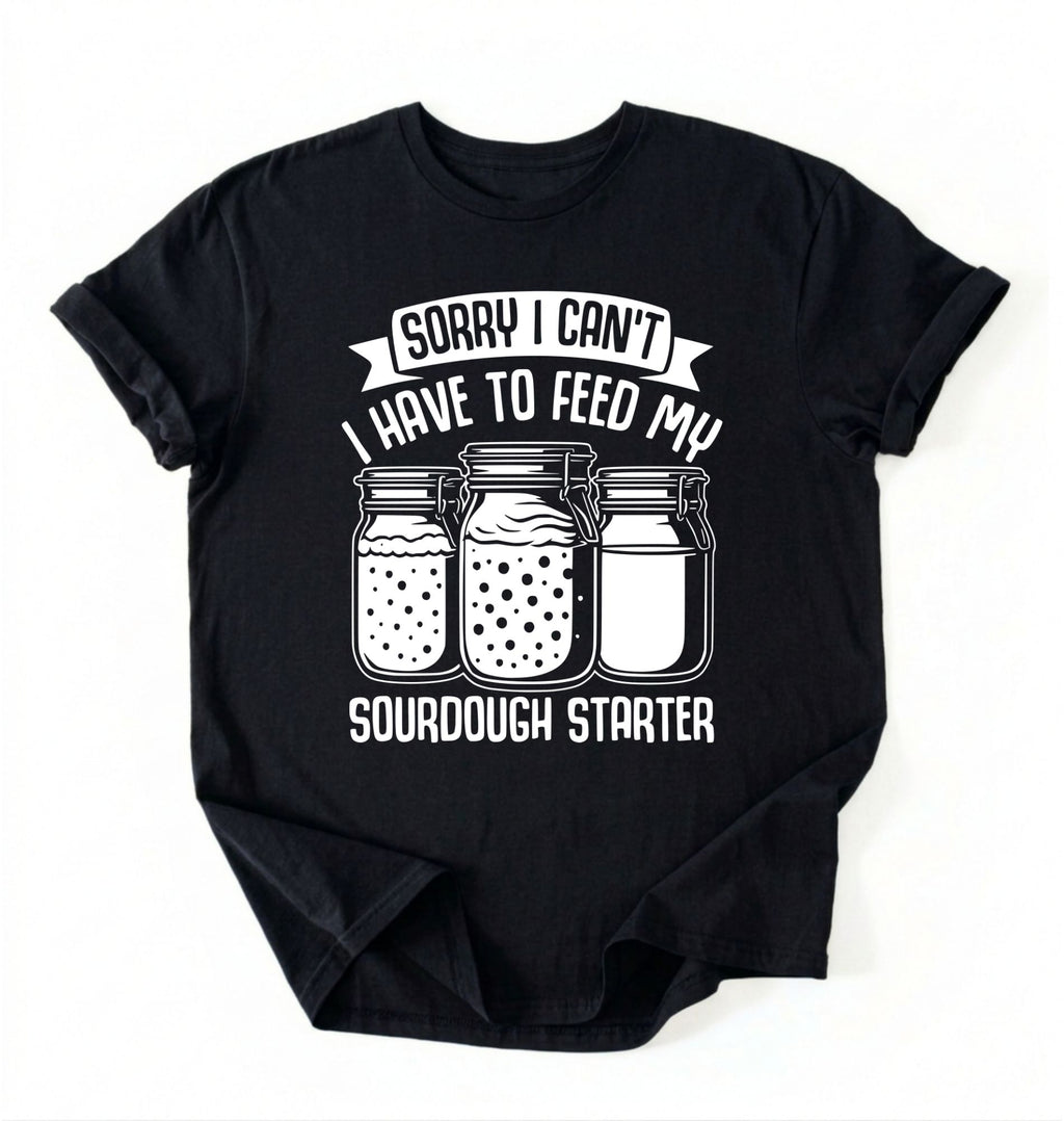 Flat lay of a black Sorry I Can't I Have to Feed My Sourdough Starter shirt with starter jar graphic on charcoal t-shirt on a white background 