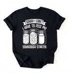 Flat lay of a black Sorry I Can't I Have to Feed My Sourdough Starter shirt with starter jar graphic on charcoal t-shirt on a white background 