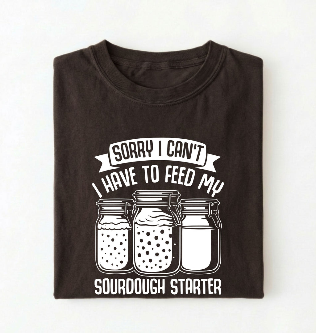 Flat lay of a brown Sorry I Can't I Have to Feed My Sourdough Starter shirt with starter jar graphic on charcoal t-shirt on a white background 