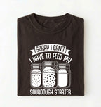 Flat lay of a brown Sorry I Can't I Have to Feed My Sourdough Starter shirt with starter jar graphic on charcoal t-shirt on a white background 
