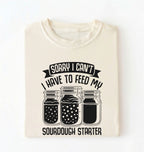Flat lay of a cream Sorry I Can't I Have to Feed My Sourdough Starter shirt with starter jar graphic on charcoal t-shirt on a white background 