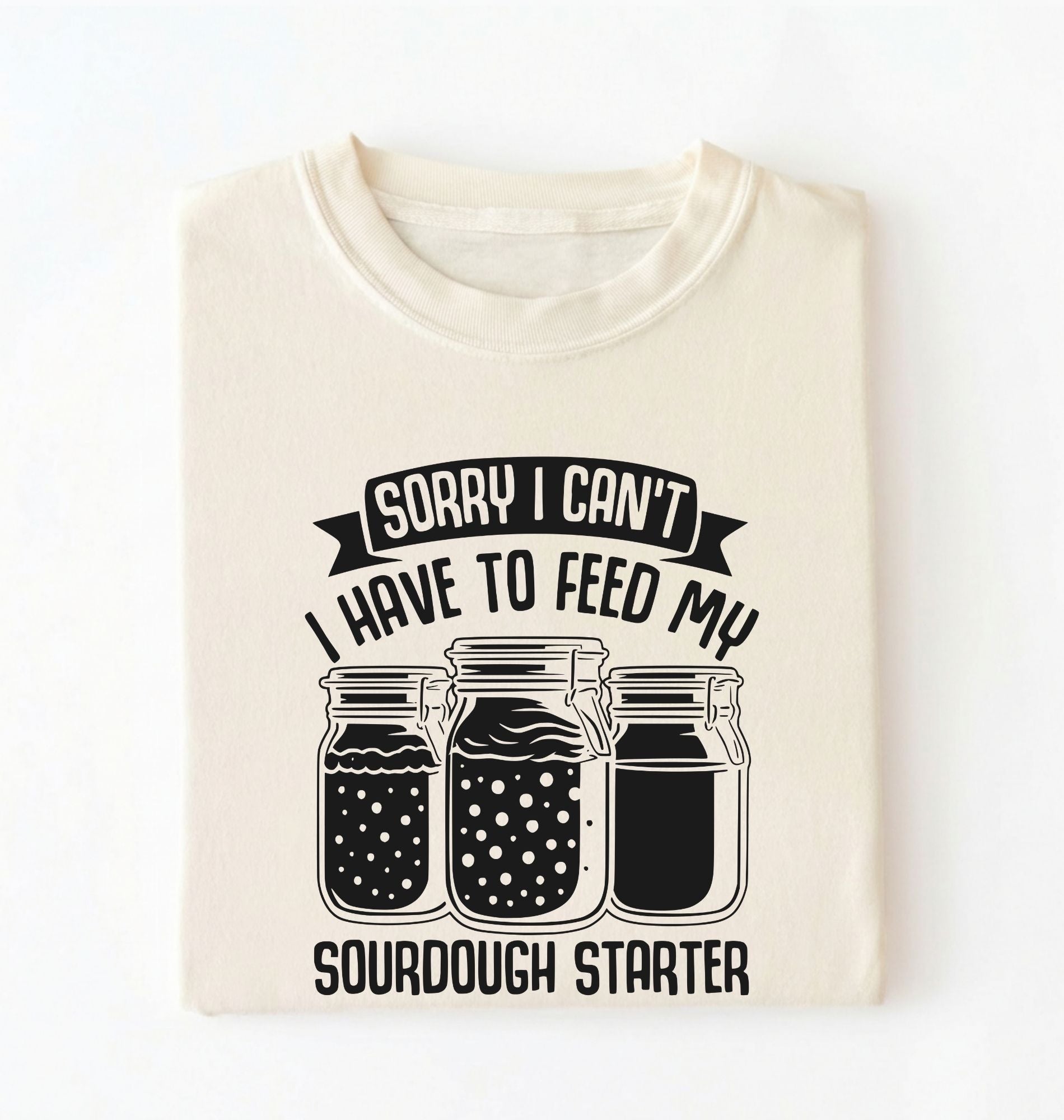 Flat lay of a cream Sorry I Can't I Have to Feed My Sourdough Starter shirt with starter jar graphic on charcoal t-shirt on a white background 