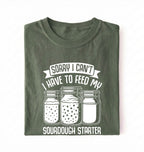 Flat lay of a green Sorry I Can't I Have to Feed My Sourdough Starter shirt with starter jar graphic on charcoal t-shirt on a white background 